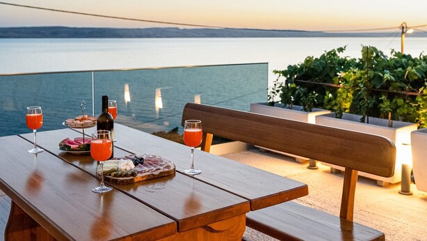Una mesa de madera con una botella, vasos y una tabla de fiambres en un balcón con vista al agua.