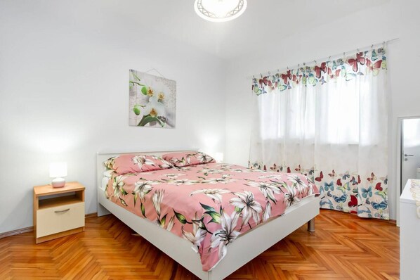 A bedroom with a bed, a nightstand, a wall-mounted lamp, a window with sheer curtains, and a floral print bedspread. - Apartments Alaga - C (Tribunj)