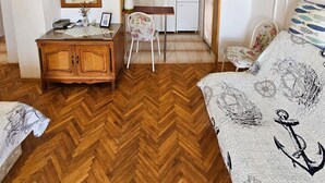 Een kamer met een houten visgraatvloer, een bed met een marineblauw beddegoed, een houten dressoir, een stoel, een bureau, een spiegel, een televisie die aan de muur is bevestigd, en een keuken in de achtergrond.