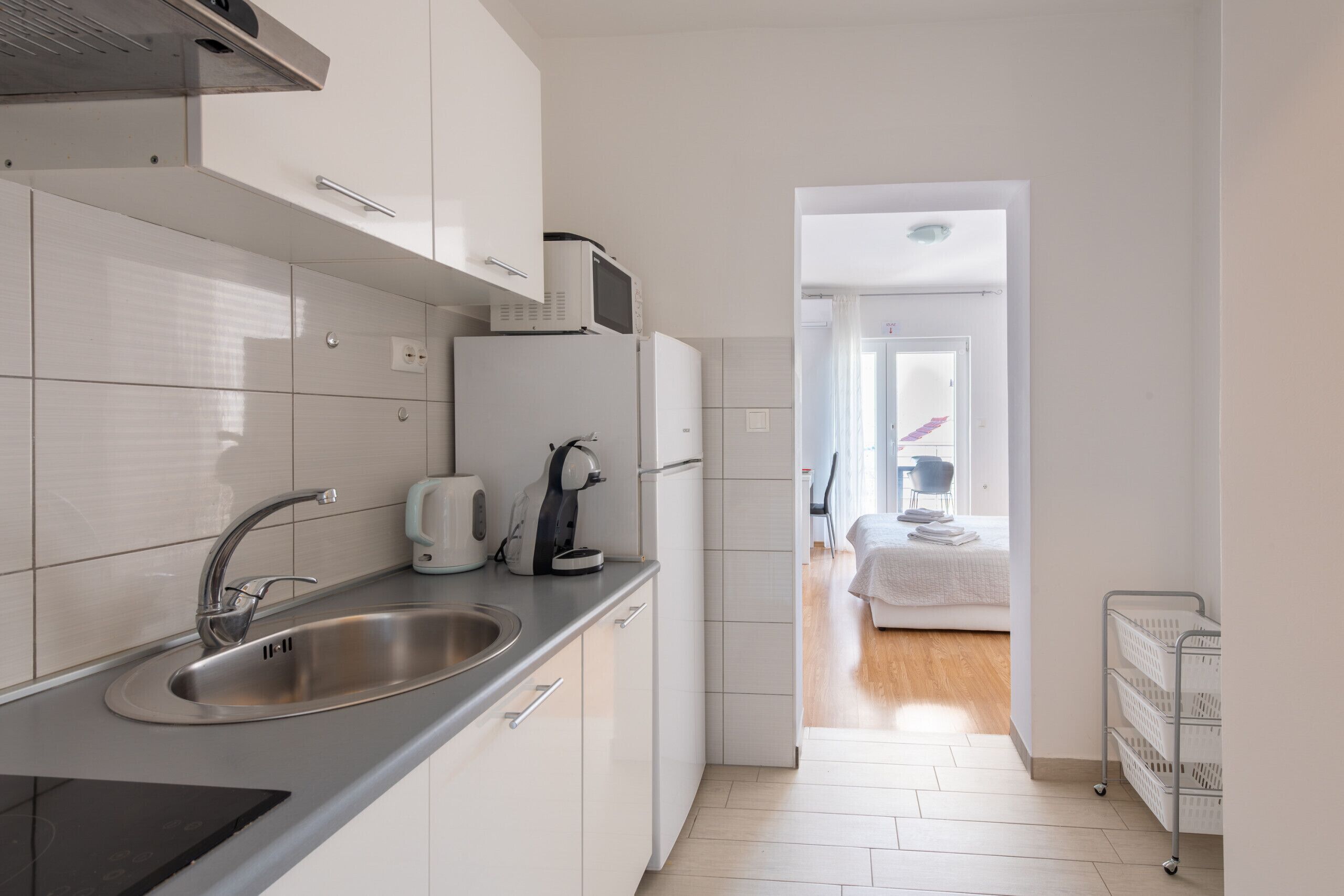 Een moderne keuken met een roestvrijstalen spoelbak, waterkoker en magnetron. Een gang leidt naar een slaapkamer met een bed en nachtkastje.