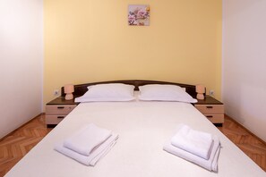 Una cama bien hecha con almohadas y toallas dobladas, mesitas de noche de madera con lámparas, y una pared con un cuadro floral.