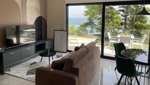 A modern living room with a sofa, a TV, a dining table with chairs, and a view of the outdoors. - Sea View Apartments Marvi (left) (Ugljan)