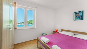 A bedroom with a bed, a nightstand, a lamp, a window with a view of the sea, and a painting on the wall.