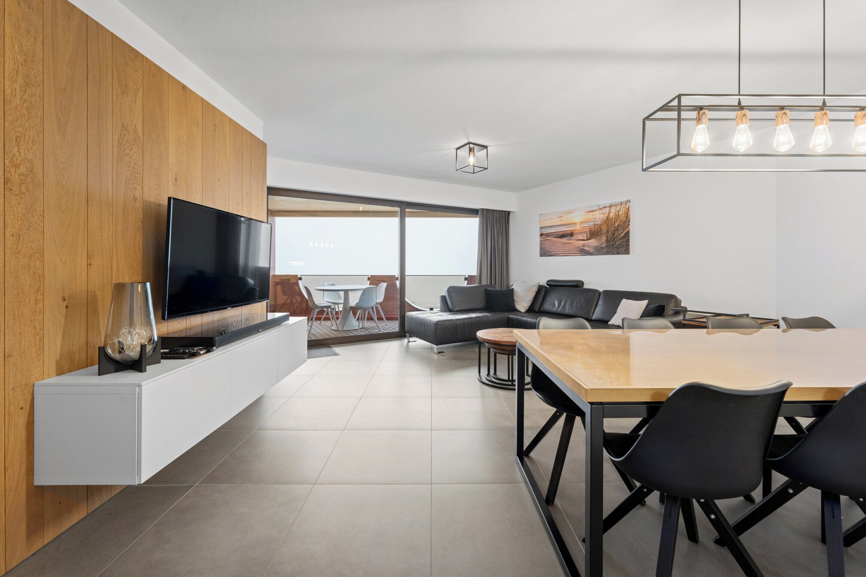 A modern living room with a flat-screen TV, wooden panel wall, and a dining area with a wooden table and black chairs.