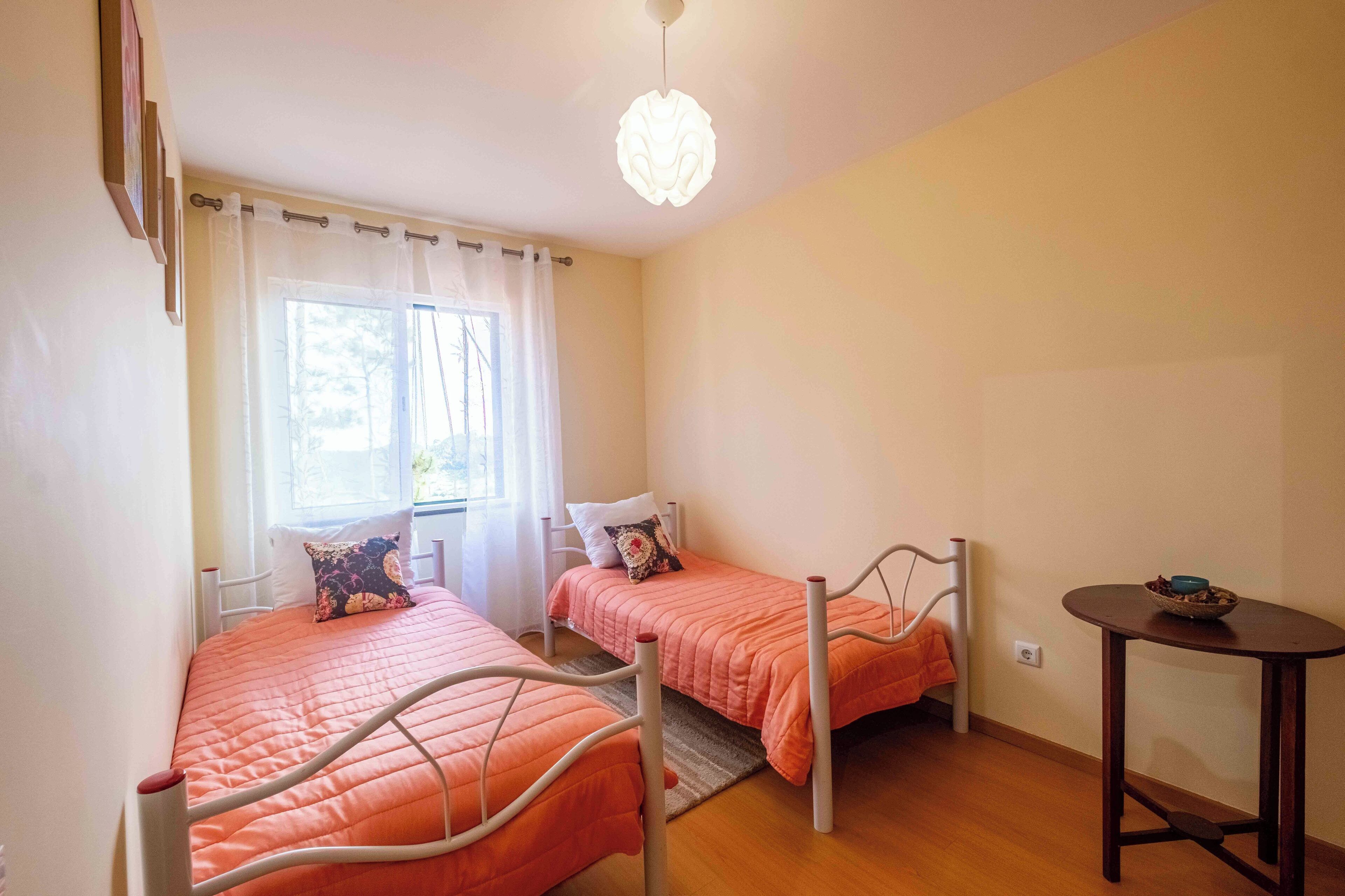 A room with two single beds, a small table, a window with curtains, and a ceiling light fixture.