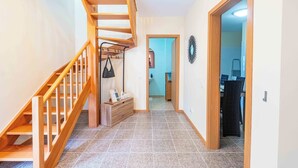 Una scala con ringhiere in legno conduce a un secondo piano. Una panca di legno con un cartello e una borsa appesa al muro. Un'porta dà accesso a un bagno con una parete blu e un lavandino.