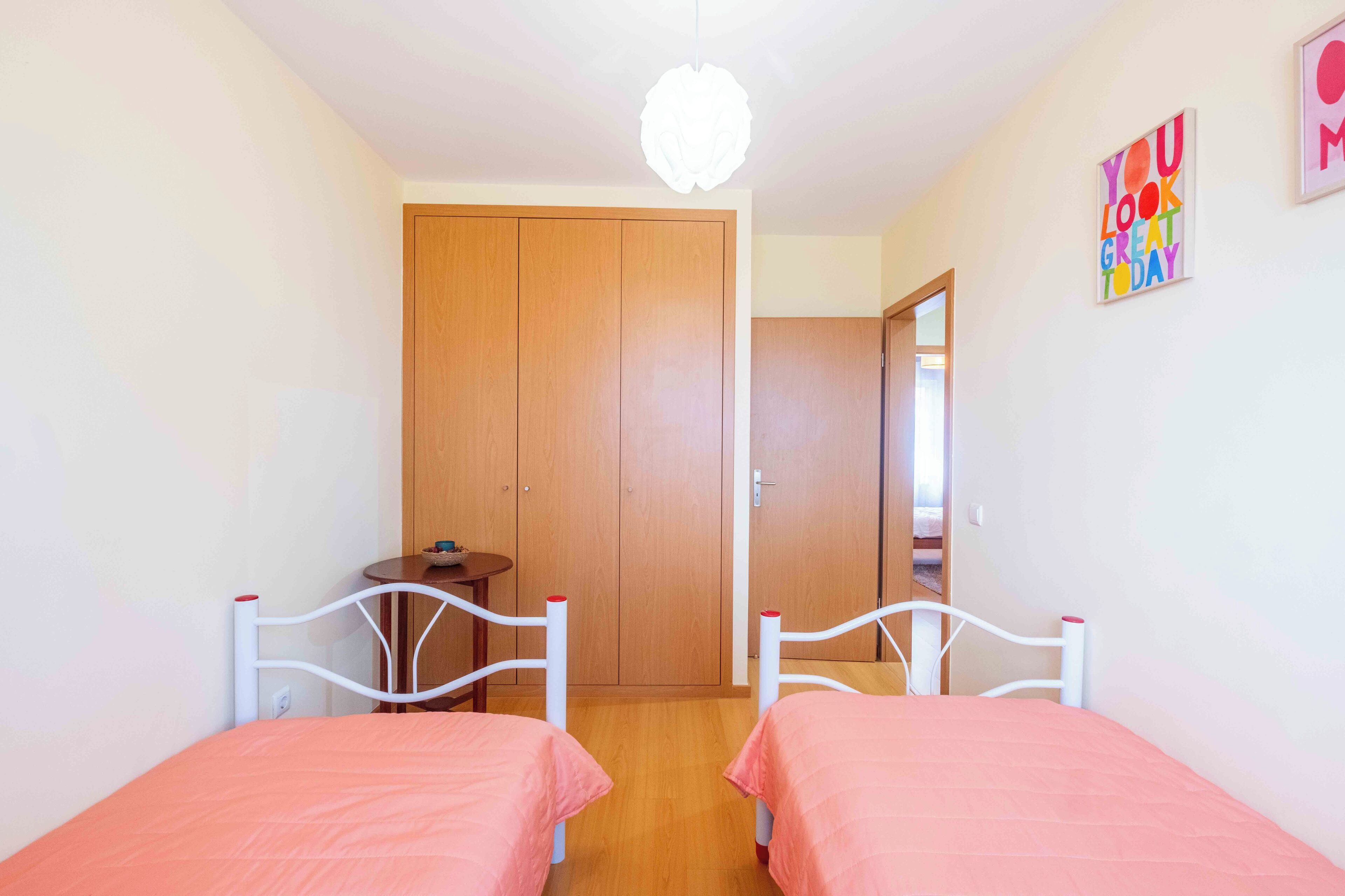 A bedroom with two single beds, a wooden wardrobe, a small table, and a ceiling light fixture.