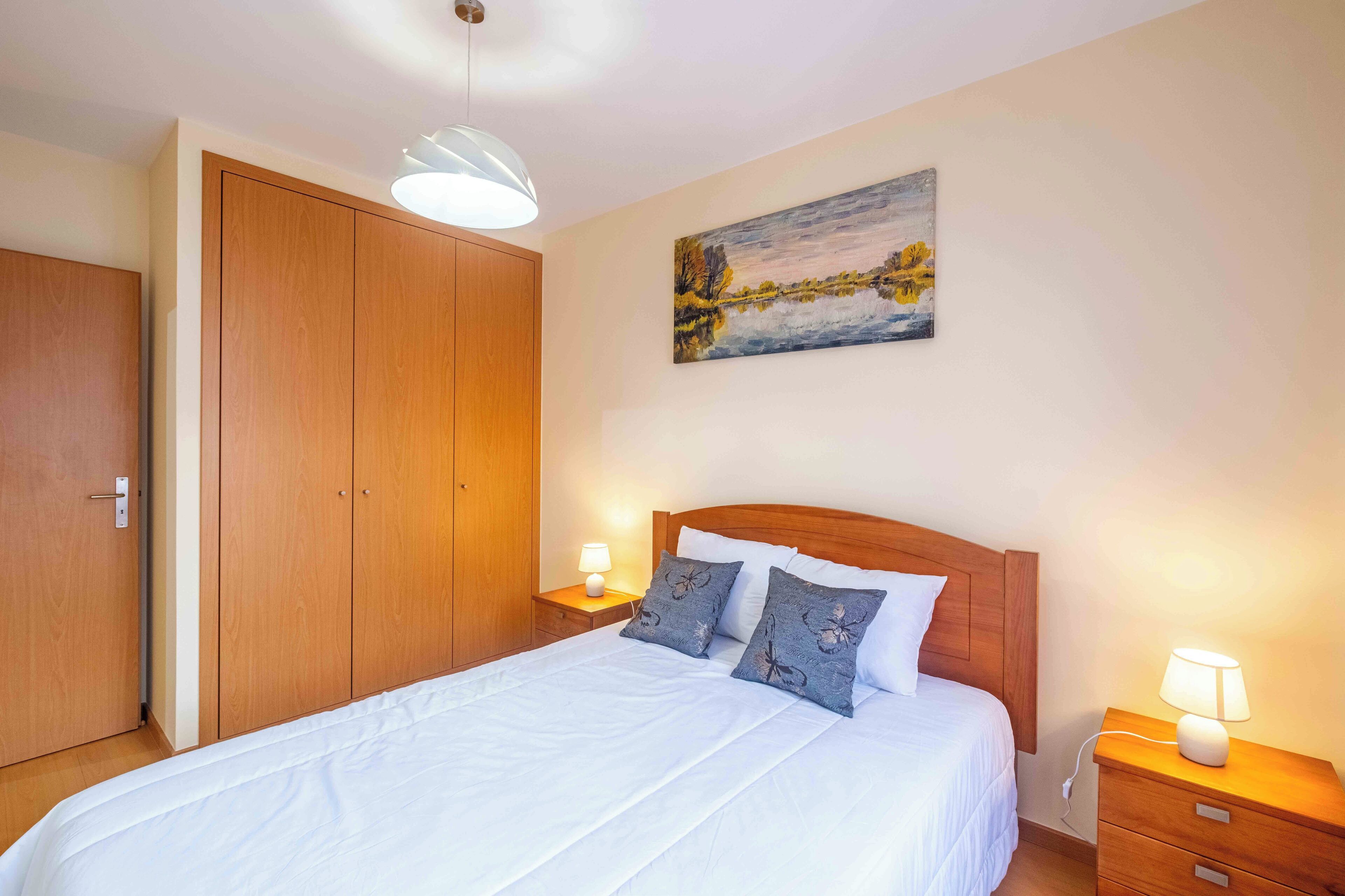 Une chambre avec une armoire en bois, un lit avec une literie blanche, deux tables de chevet avec des lampes et un tableau au mur.