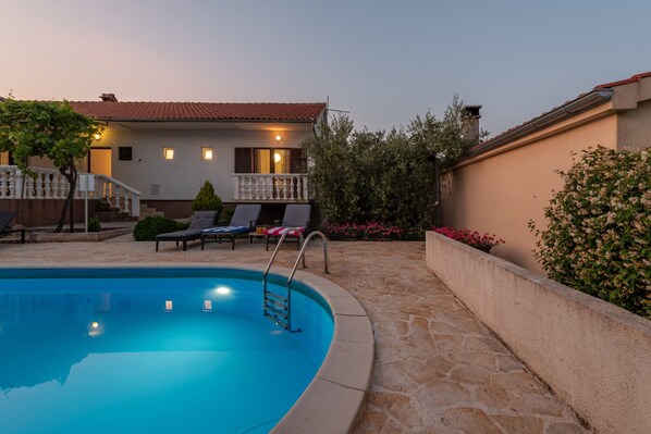 A swimming pool with a stone deck, a house with a balcony, and a garden with flowers.