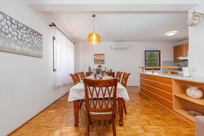 A dining area with a wooden table, chairs, a hanging light fixture, and a kitchen in the background.