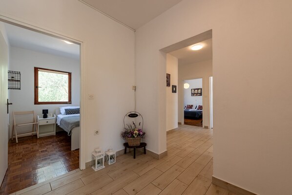 A hallway with wooden flooring, a bed with a white headboard, a small table with a basket of flowers, and a white cabinet.