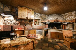 A rustic kitchen with a stone wall, wooden cabinets, a built-in oven, and a dining area with a wooden table. - Villa Smilje ZadarVillas (Slivnica)