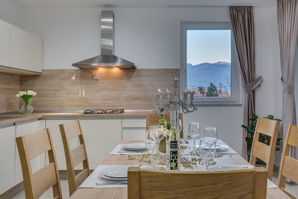 Un comedor con una mesa lista para cuatro personas, una cocina moderna con extractor de acero inoxidable y una ventana con vistas a montañas.