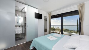 A modern hotel room with a large bed, a TV, and a balcony with a view of the sea.
