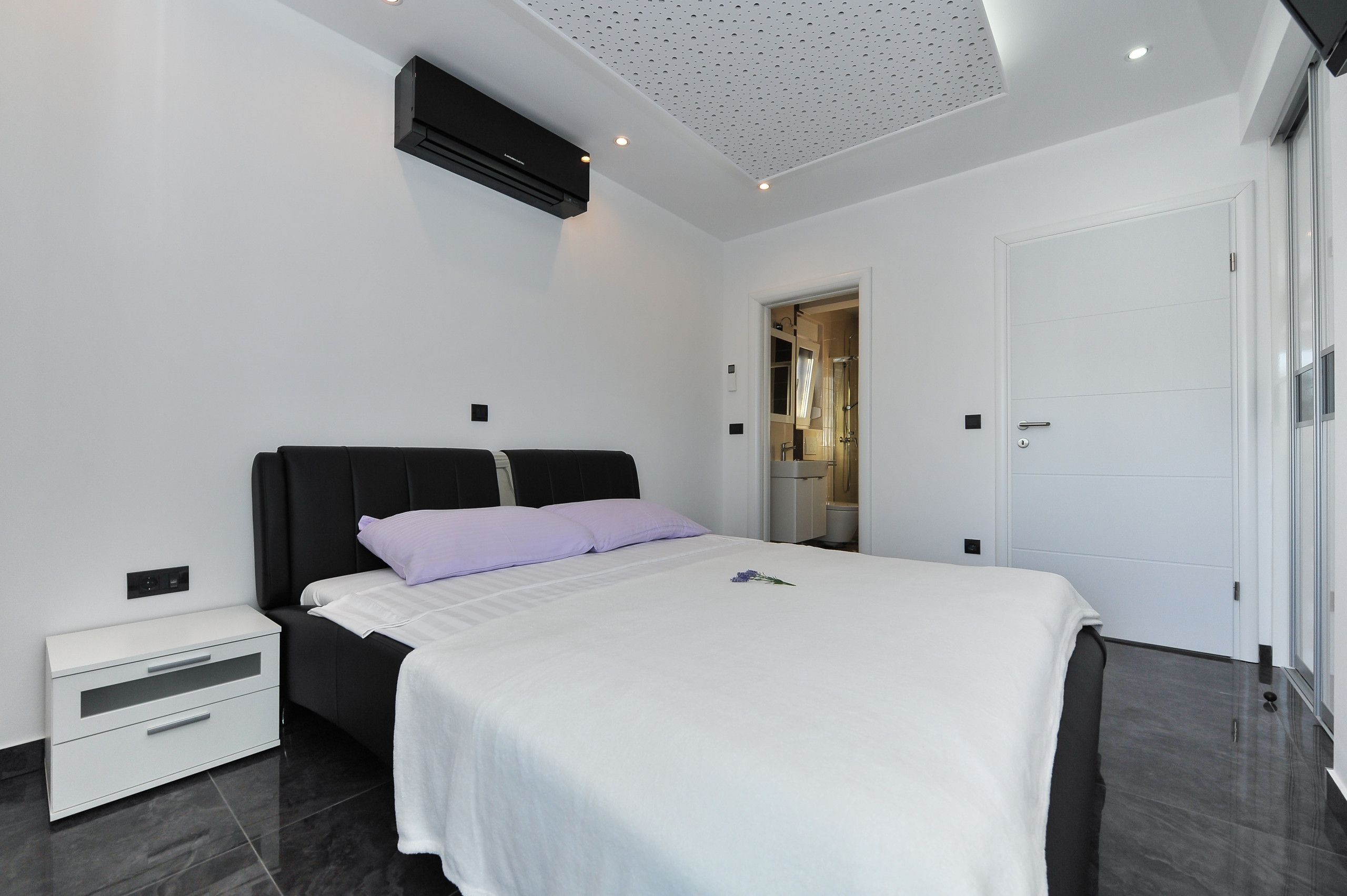 A modern bedroom with a black headboard, white bedding, a white nightstand, and a ceiling with recessed lighting.