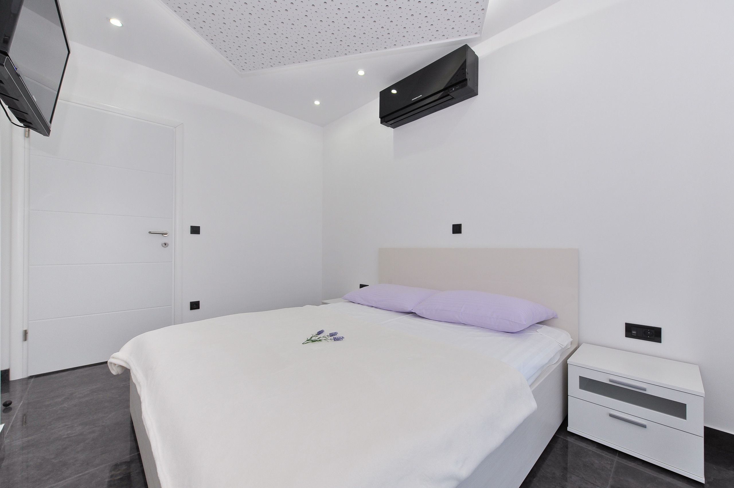 A modern bedroom with a bed, a nightstand, a TV mounted on the wall, and a ceiling with recessed lighting.