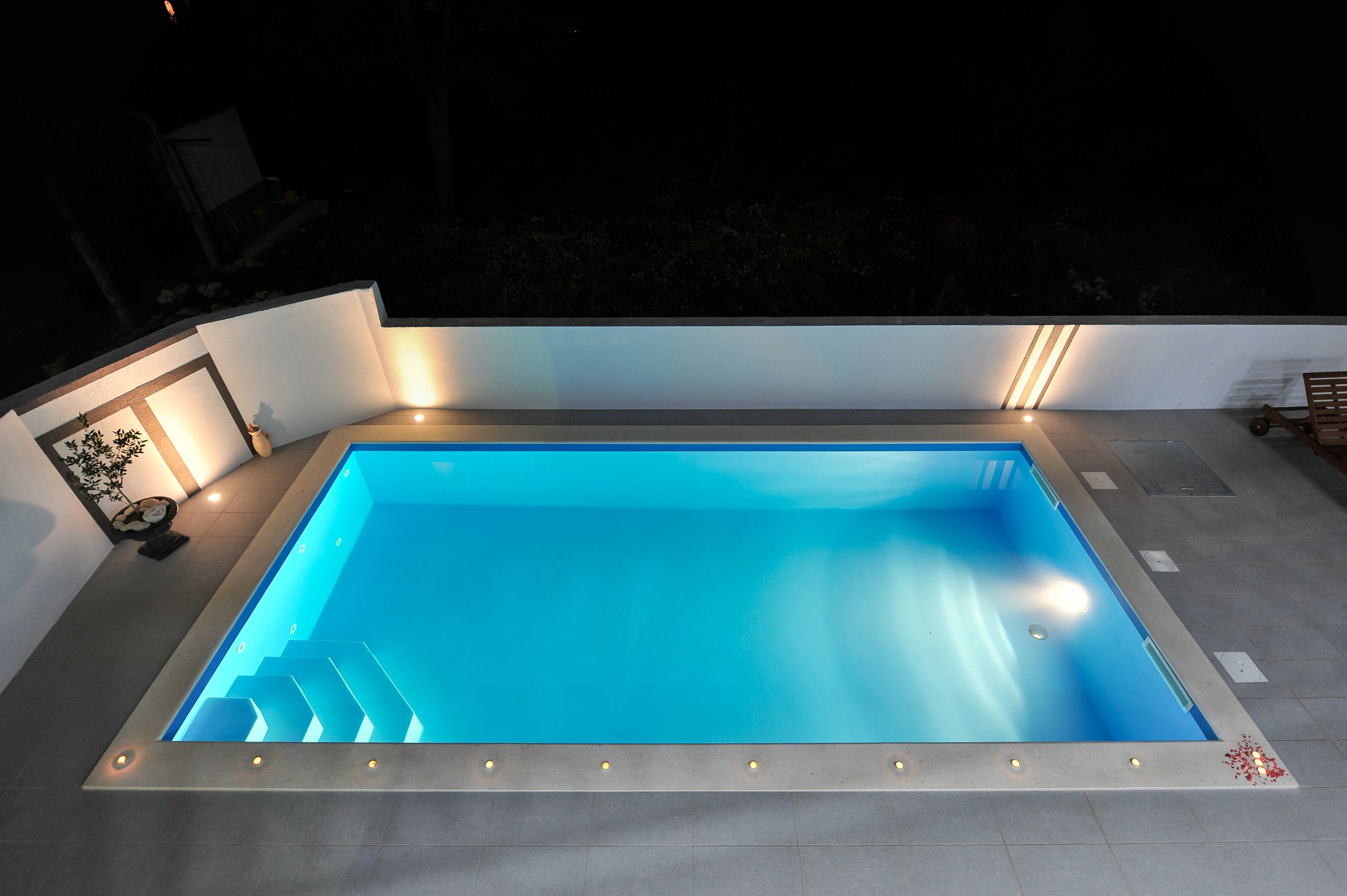 A rectangular swimming pool with a clear blue interior, surrounded by a tiled deck and illuminated by underfloor lighting.