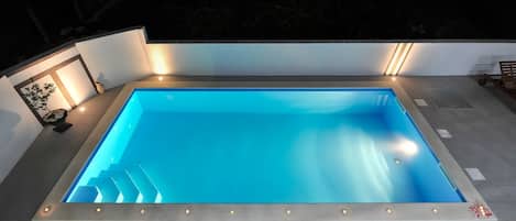 A rectangular swimming pool with a clear blue interior, surrounded by a tiled deck and illuminated by underfloor lighting.