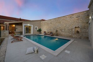 A swimming pool with stone walls, a patio with lounge chairs, and a door leading to a covered area.