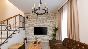 A living room with a stone wall, a flat-screen TV, a wooden coffee table with fruit and a bottle, a brown sofa, and a staircase with black railings. - Villa Imperia ZadarVillas (Pridraga)