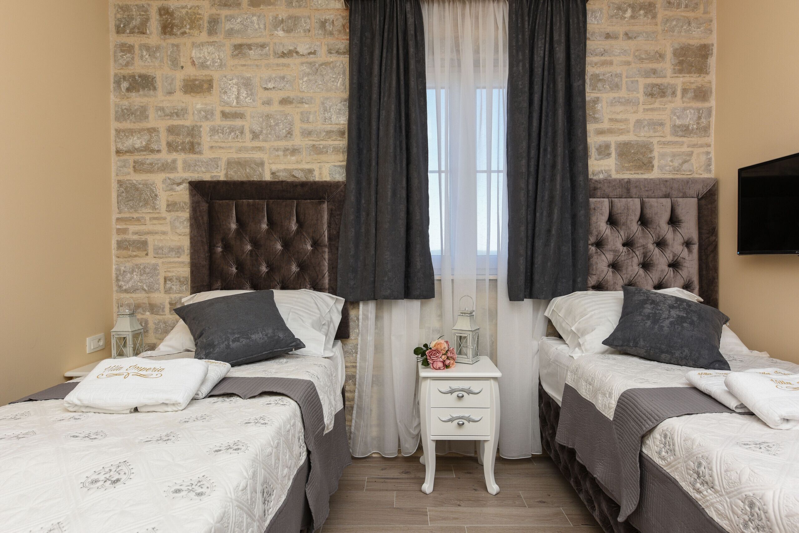 Ein Zimmer mit zwei Betten, einer Steinwand, einem Fenster mit Vorhängen sowie einem kleinen Nachttisch mit Lampe und Blumen.