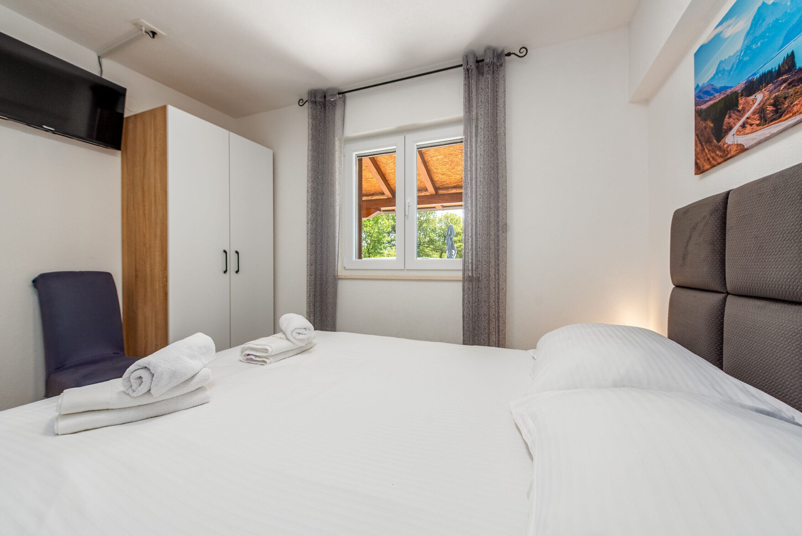 Une chambre d’hôtel avec un lit, une chaise, une télévision, une fenêtre avec des rideaux et un tableau au mur.