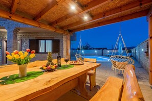 A wooden dining table with a vase of tulips, a fruit bowl, and a wine glass. There are wooden chairs and a hanging chair near a pool. - Villa Kamen ZadarVillas (Pridraga)