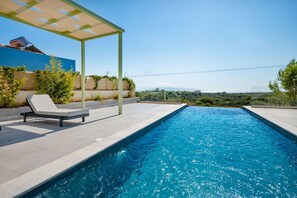 Una piscina con un pergolato e poltrone prendisole, immersa nel verde e con vista panoramica sul paesaggio.