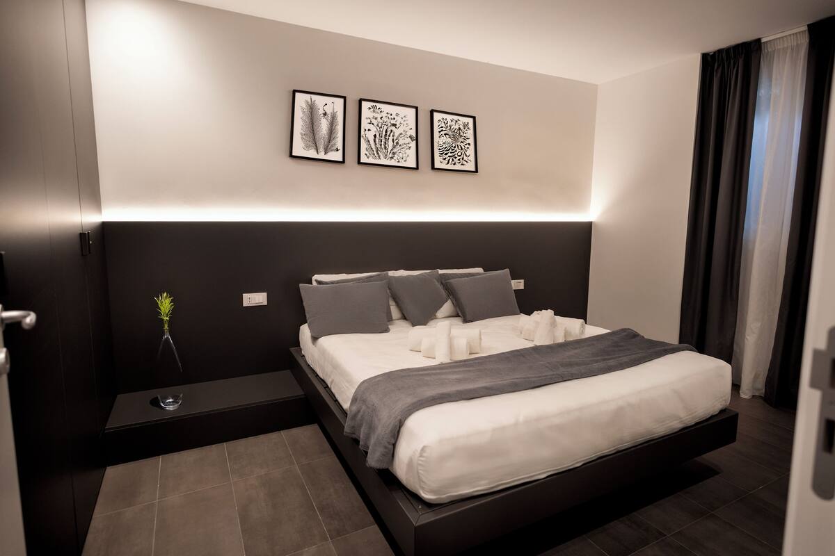 Une chambre à coucher moderne avec un grand lit, une table de chevet et trois reproductions botaniques encadrées accrochées au mur.