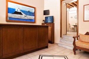 Uma área de recepção de hotel com um balcão de madeira, um quadro emoldurado retratando uma paisagem costeira, uma televisão e uma escadaria para outro espaço.