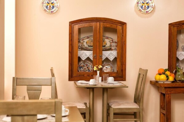 A dining area with a wooden cabinet displaying decorative plates, a table set for two with chairs, and a vase with flowers on the table. - Egli Hotel (Andros)