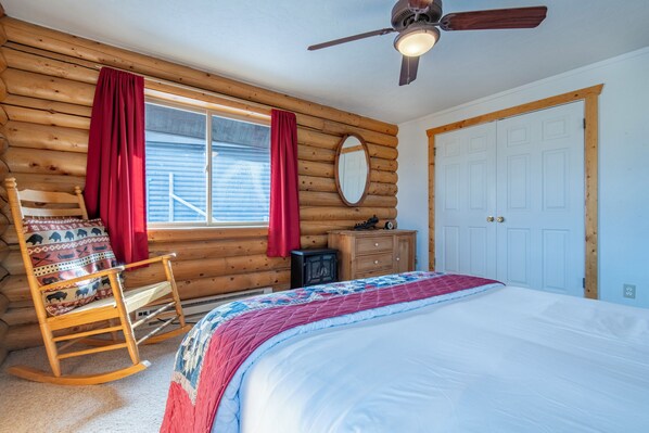 A log cabin bedroom with a bed, rocking chair, wooden walls, and a ceiling fan.