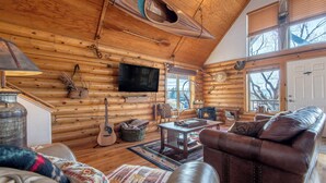 Interior de una cabaña de troncos con techo de madera, chimenea, televisor montado en la pared y sofá de cuero.