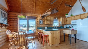 Interior de una cabaña de troncos con una mesa de comedor de madera, sillas, una cocina con armarios de madera, una isla grande y lámparas colgantes.
