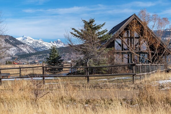 A rustic wooden cabin with a steep roof, surrounded by a wooden fence and a snow-covered landscape with mountains in the background. - Cozy Teton Cabin (Victor)
