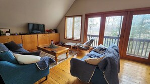 A cozy living room with a wooden floor, a sofa, a coffee table, and a TV. There are large windows and a balcony.