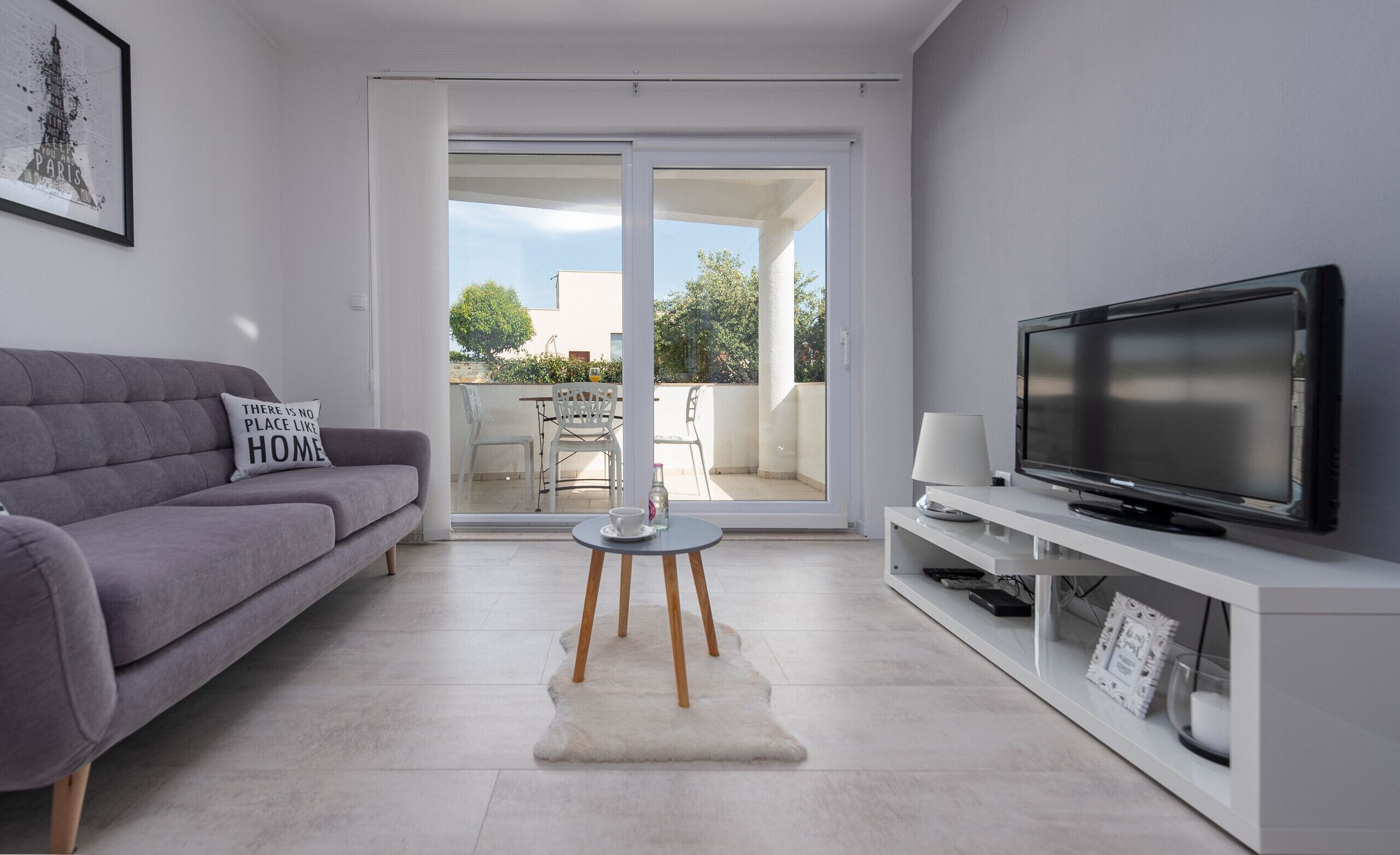 Un salon moderne avec un canapé gris, un téléviseur à écran plat posé sur un meuble blanc et une porte vitrée donnant sur une terrasse extérieure.
