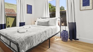 Ein ordentlich bezogenes Bett mit weißen Laken und einer grauen Decke, ein Nachttisch mit einer Lampe und ein Fenster mit Vorhängen.