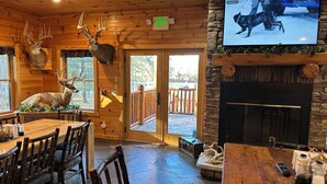 Un intérieur rustique avec un coin repas en bois, des têtes de cerf accrochées au mur, une cheminée et un téléviseur diffusant une scène hivernale enneigée.