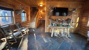 L’intérieur d’une cabane en rondins, avec un sol carrelé en pierre, des meubles en bois, des têtes de cerf accrochées aux murs, une cheminée et un escalier.