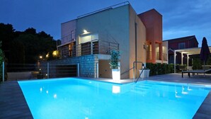A swimming pool with a modern building in the background, a covered seating area, and a stone wall. - Villa Amarena ZadarVillas (Zadar)