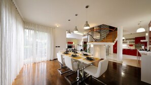 A modern dining area with a wooden floor, white dining chairs, and a table set for four. A staircase leads to a kitchen area with red cabinets. - Villa Amarena ZadarVillas (Zadar)