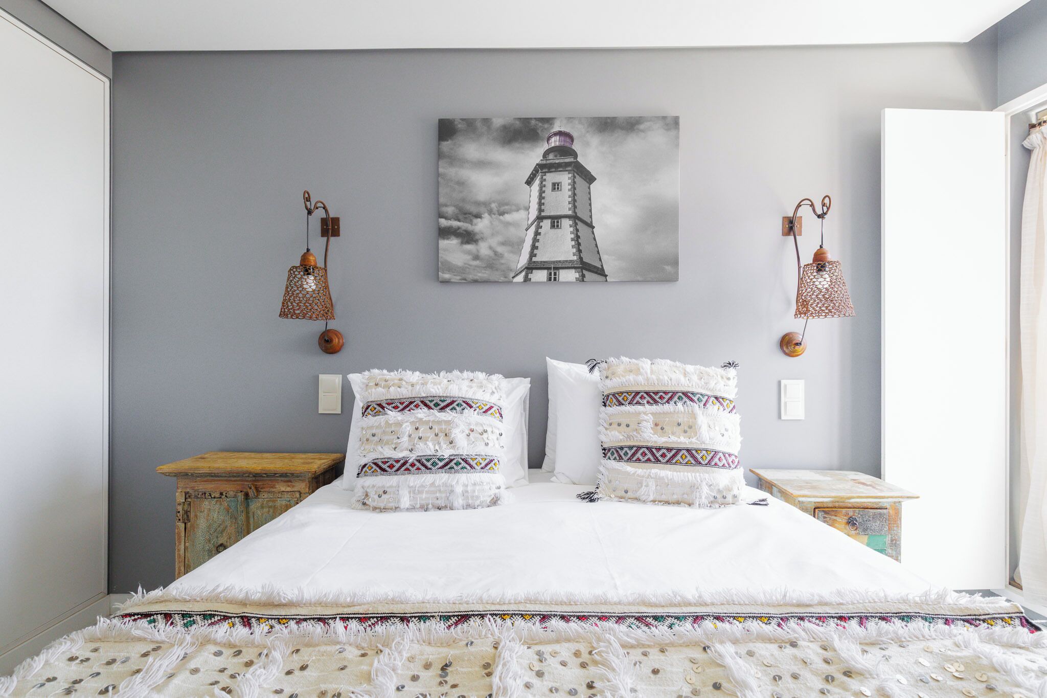 Une chambre à coucher avec un lit, deux oreillers, une table de chevet, une lampe fixée au mur et un tableau représentant un phare.