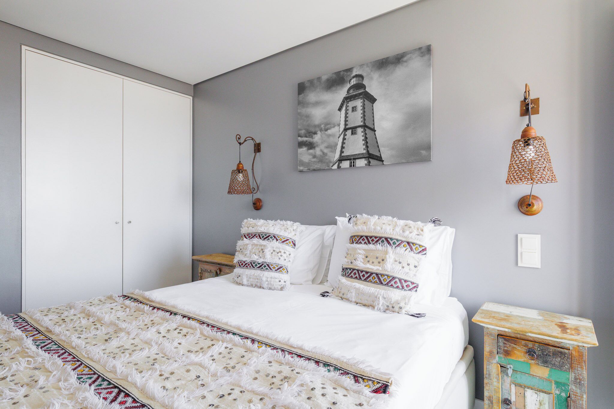 A bedroom with a bed, a painting of a lighthouse, a hanging lamp, and a small table.