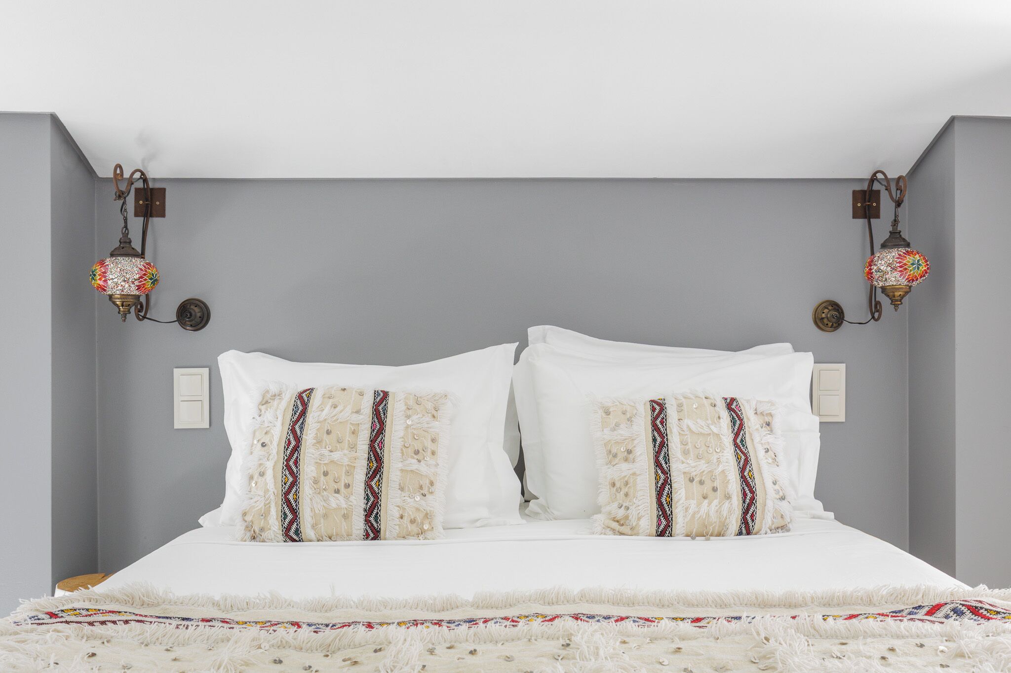Una cama con ropa de cama blanca y almohadas decorativas, dos lámparas fijadas a la pared y una pared gris lisa.