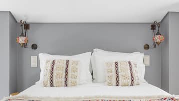 Una cama con ropa de cama blanca y almohadas decorativas, dos lámparas fijadas a la pared y una pared gris lisa.