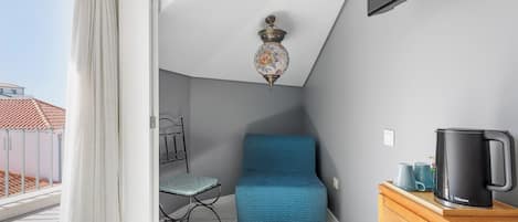 Quarto com um sofá azul, uma mesa pequena com uma cafeteira preta, uma cadeira de metal e um lustre no teto.