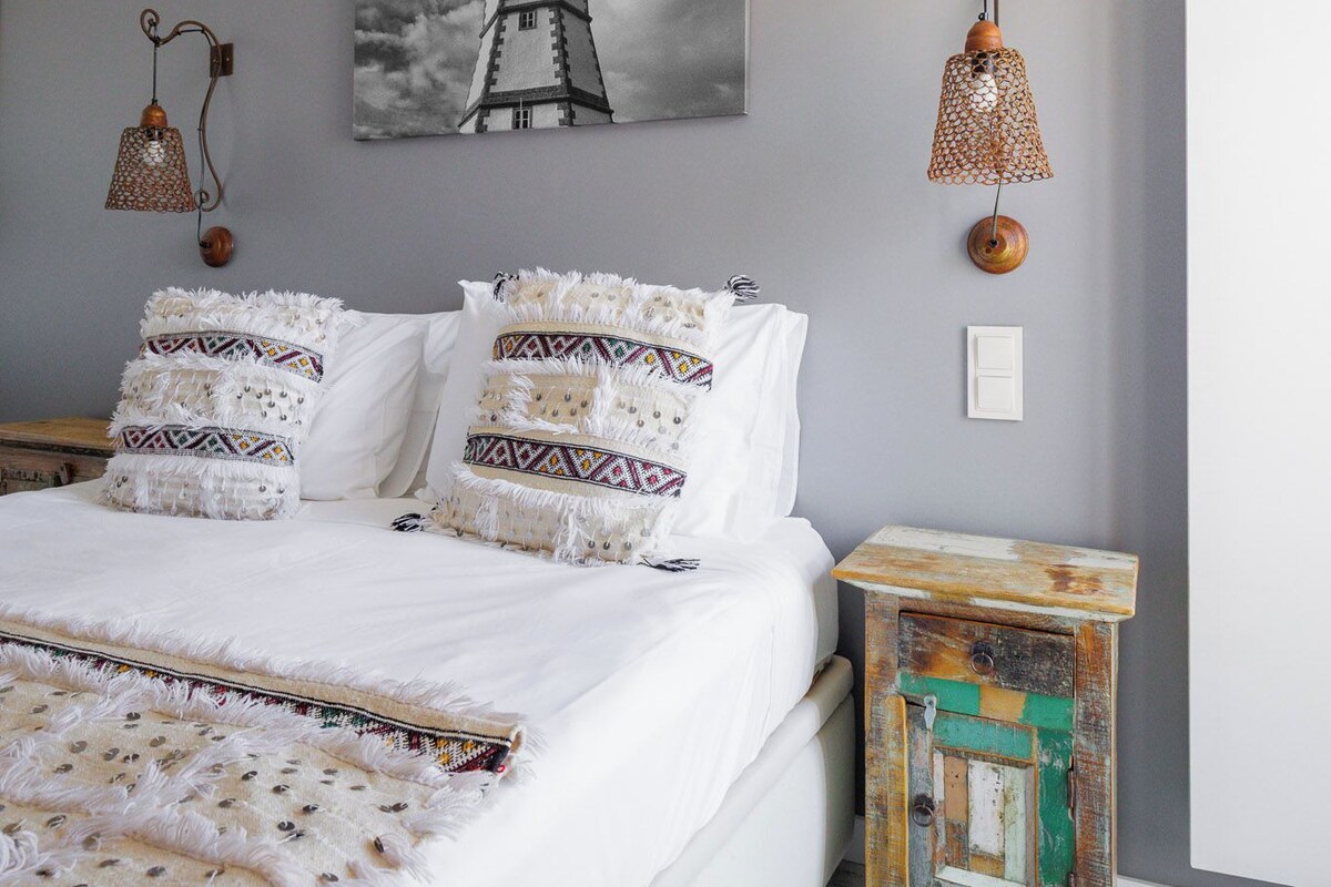 A bedroom with a bed, a wall-mounted lamp, a picture of a lighthouse, and a small wooden nightstand.