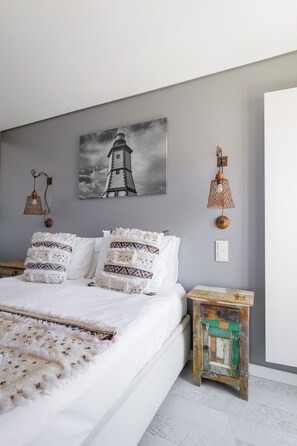 Un dormitorio con una cama, una lámpara de pared, una imagen de un faro y una mesita de noche de madera.