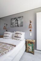 Quarto com uma cama, luminária na parede, quadro de um farol e uma pequena mesa de cabeceira de madeira.
